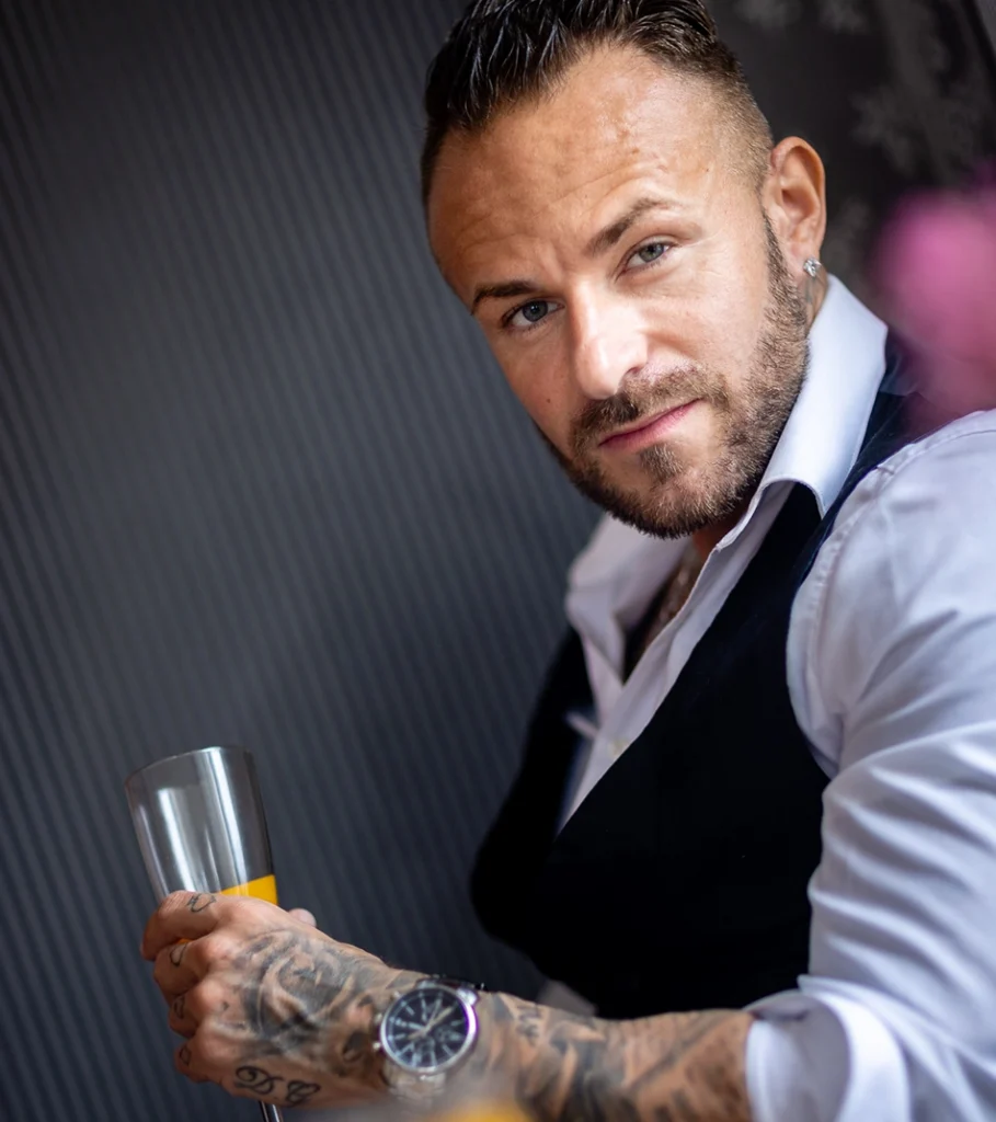 Snake Dave mit Cocktailglas an der Bar, Weste-Hemd-Kombination und sichtbaren Tattoos