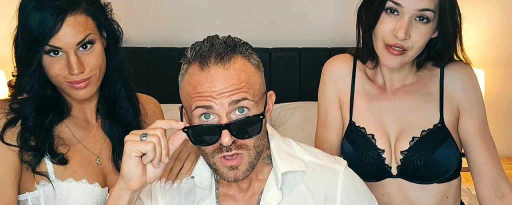 Snake Dave mit Sonnenbrille zwischen zwei jungen Frauen auf einem Bett – Querformat